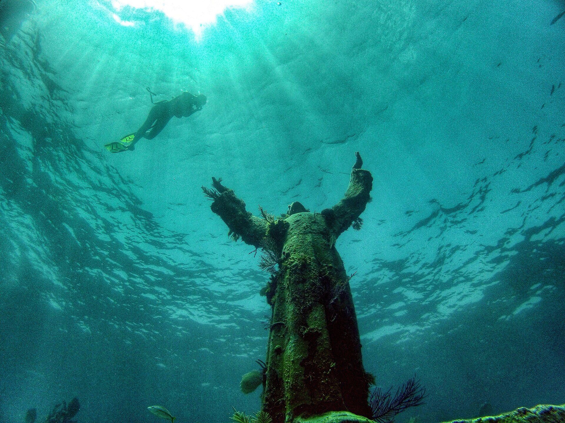 A diver swimming above the bronze Christ of the Abyss statue in blue water