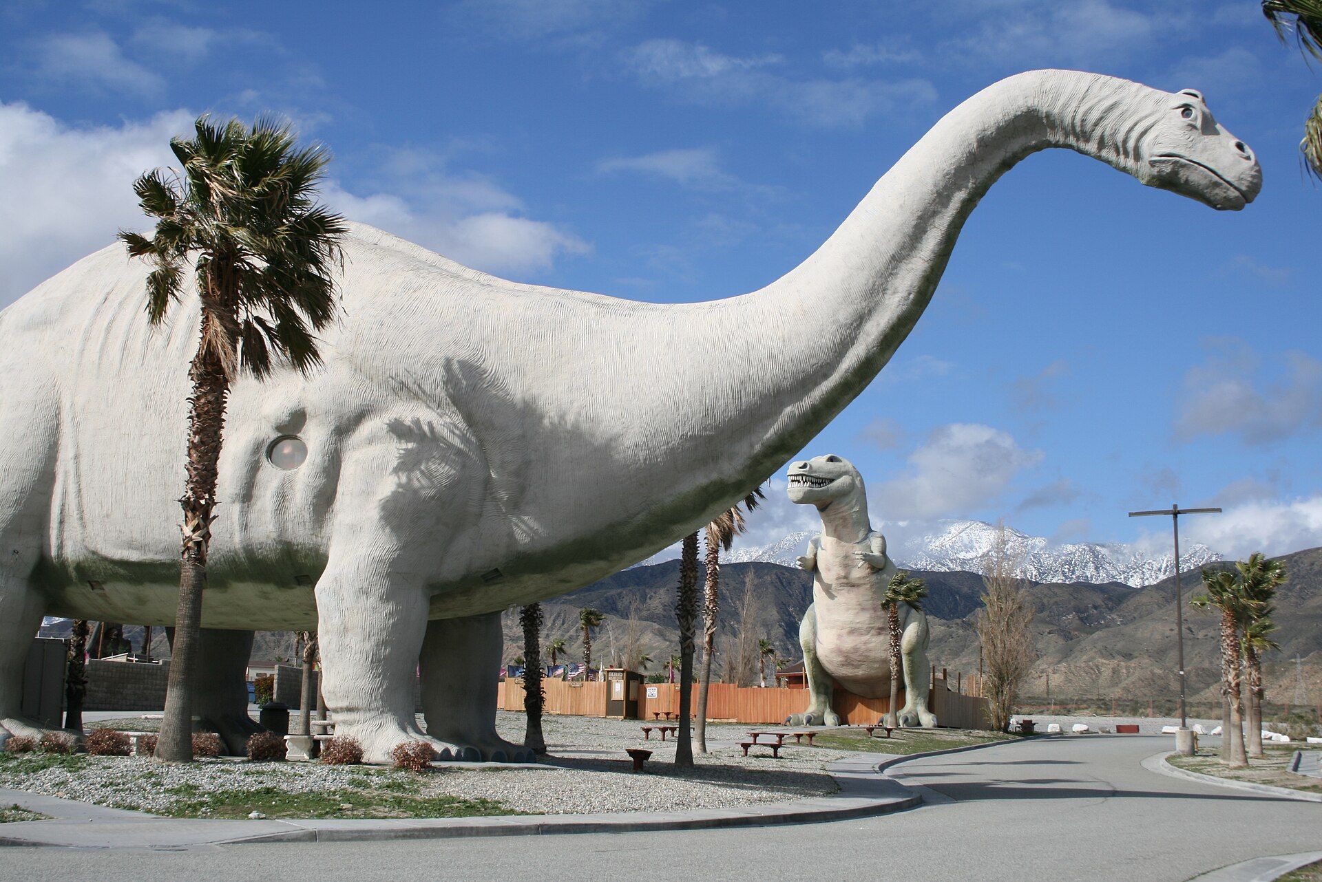 A huge grey concrete brontosaurus standing beside a desert road under palms