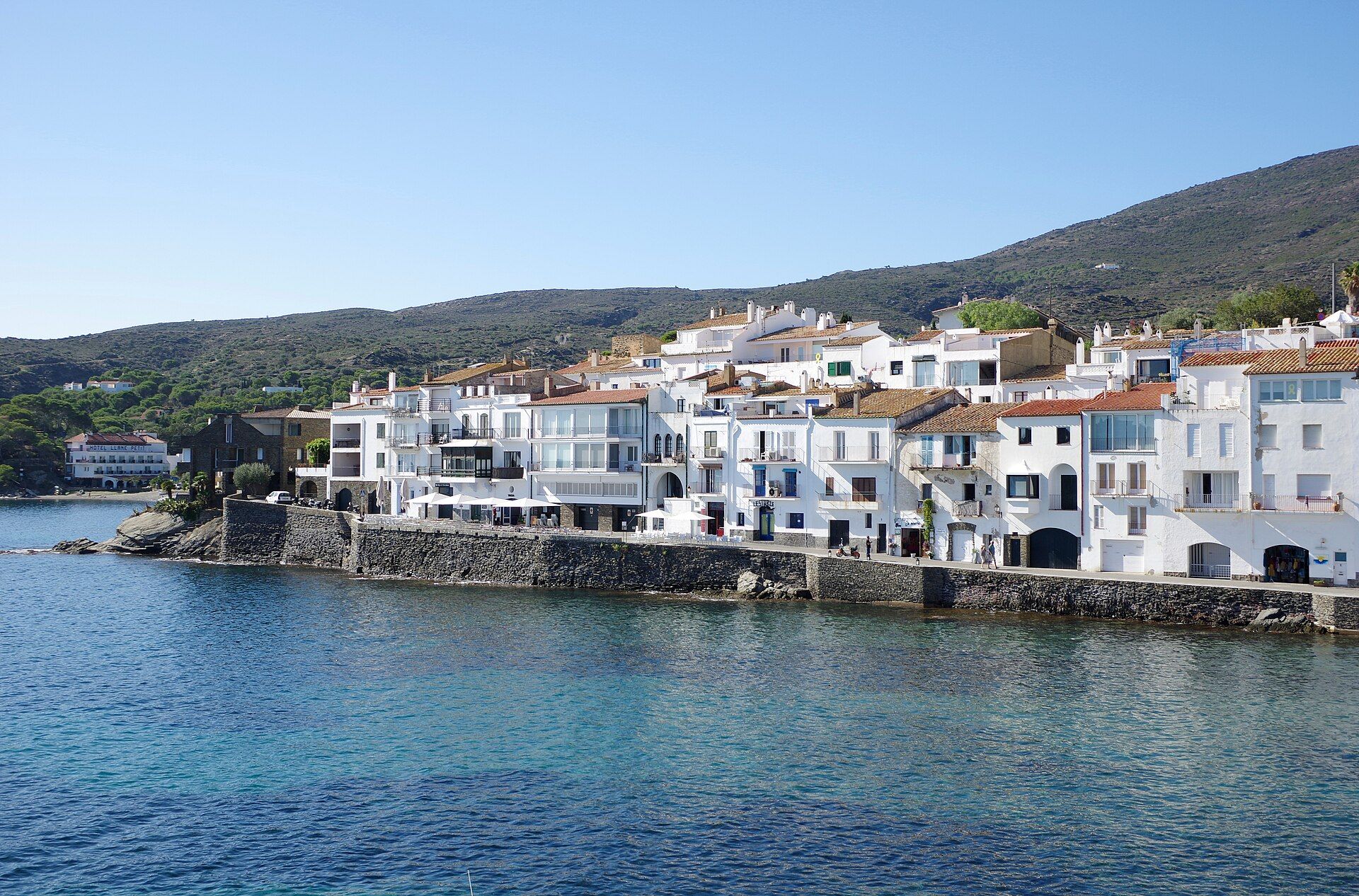 White houses with terracotta roofs along the sea wall at Cadaqués, seen across the water of the bay with scrub-covered hills behind