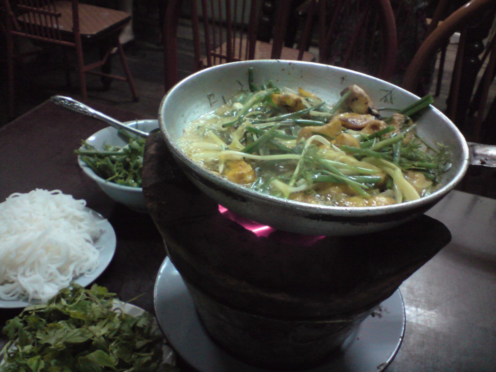 A clay pot of chả cá, fish with dill and spring onion, over a flame with noodles and herbs