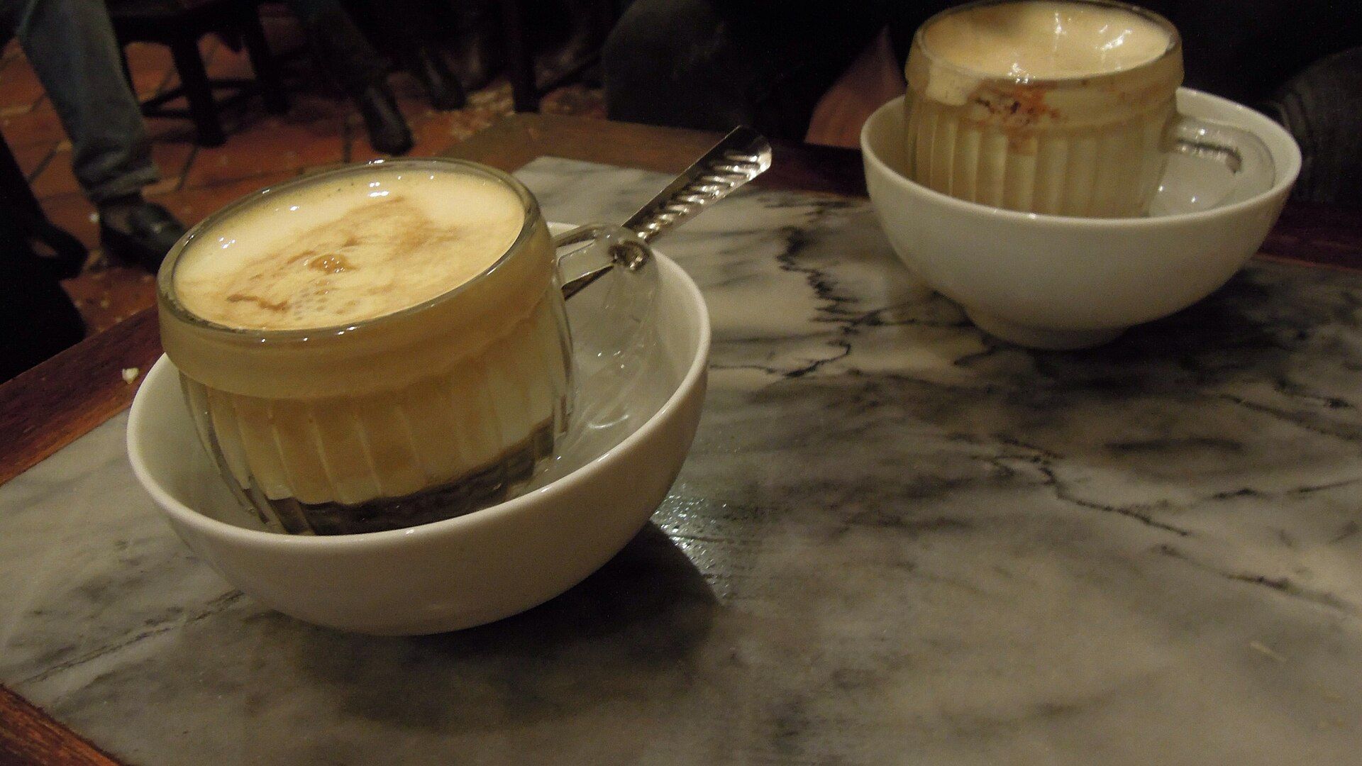 Two cups of foamy egg coffee in bowls of hot water on a marble table
