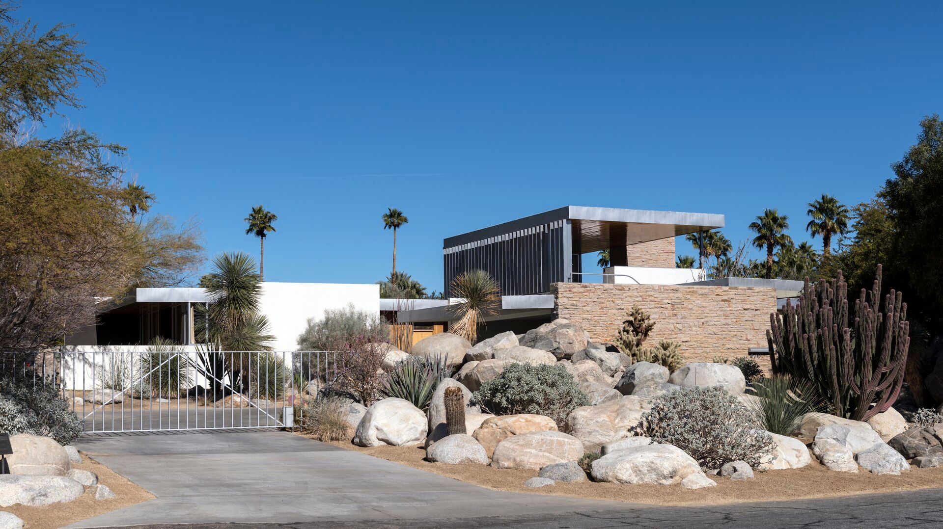 A low flat-roofed desert house behind a gate, with boulders and cactus in the front garden