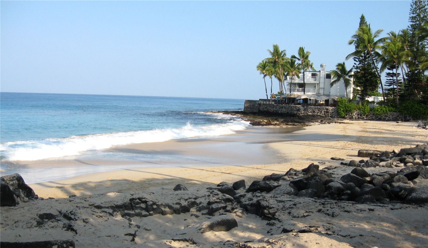Waves washing onto a small sand beach framed by black lava rock and palms