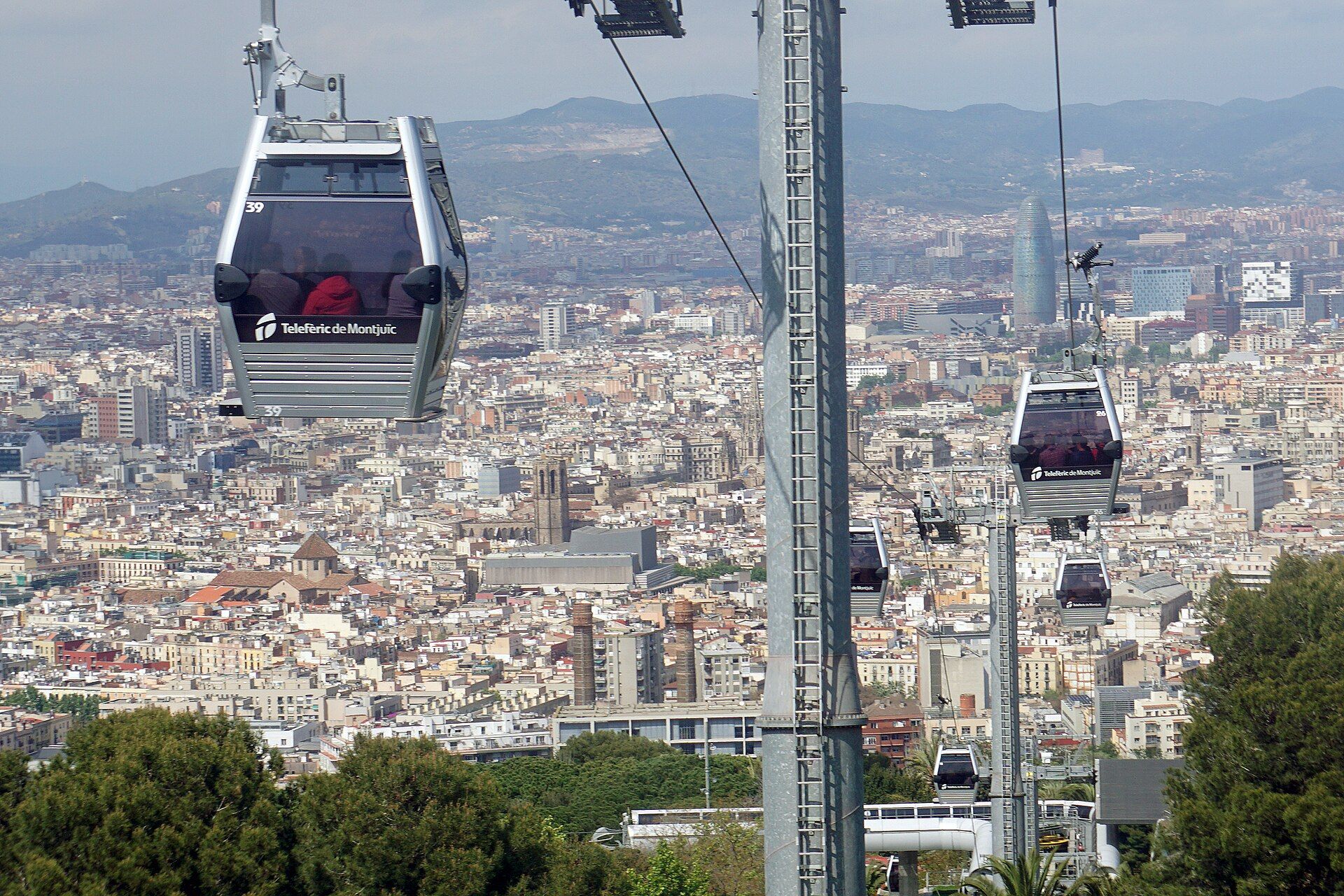 Cabins of the Montjuïc cable car passing a pylon, with the rooftops of Barcelona and the Collserola hills behind