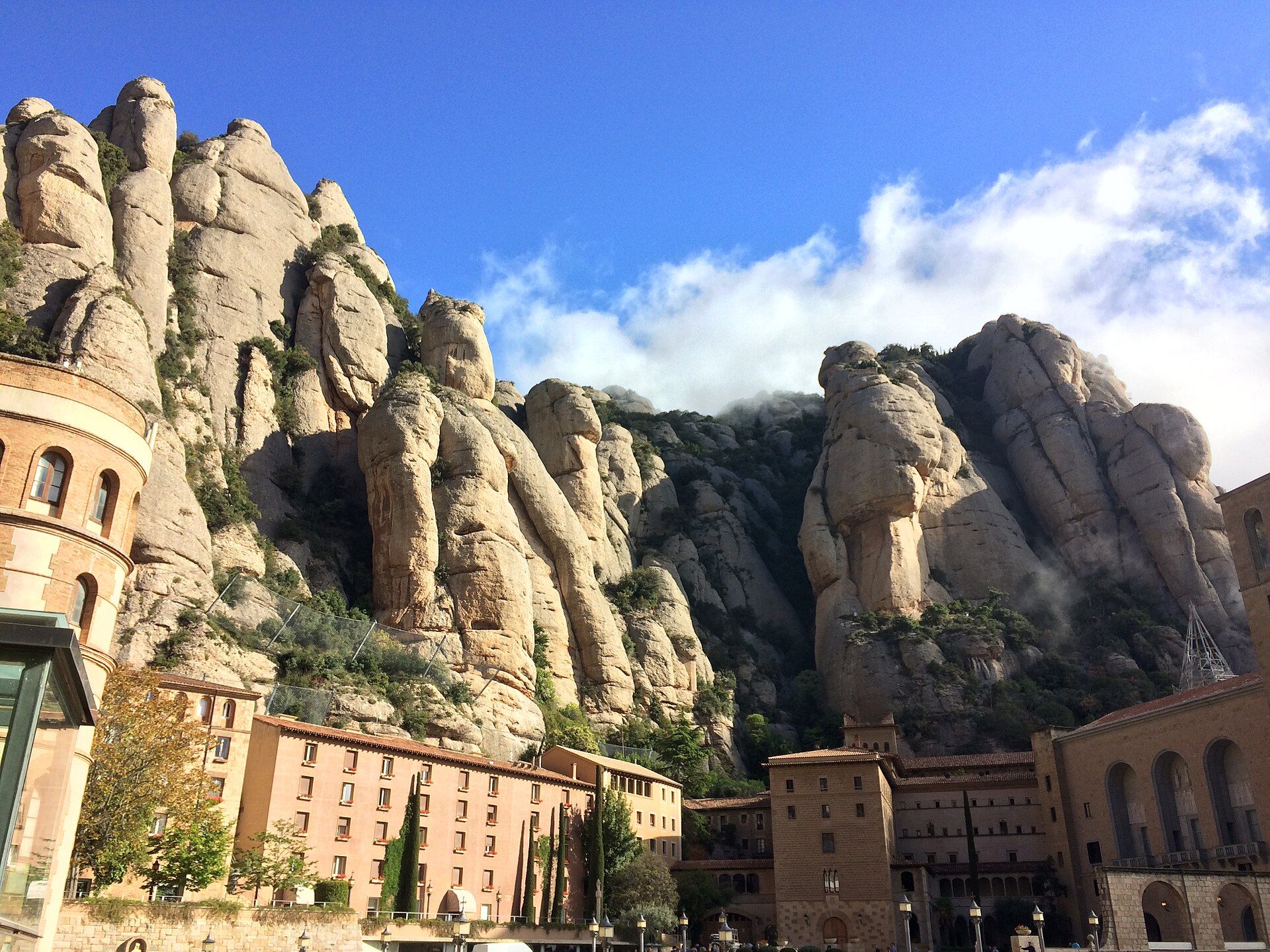 The ochre buildings of Montserrat abbey at the foot of rounded rock pillars, with cloud catching on the peaks