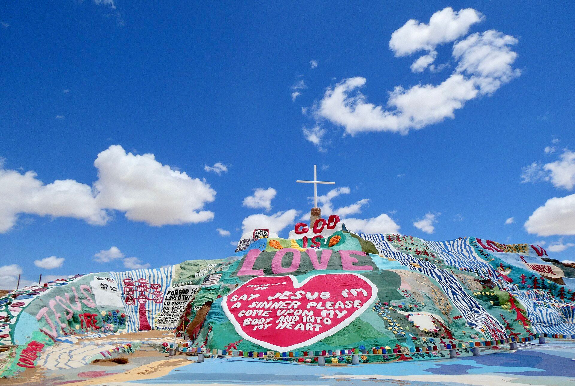 A painted adobe hillside covered in bright stripes, flowers and lettering under a blue sky