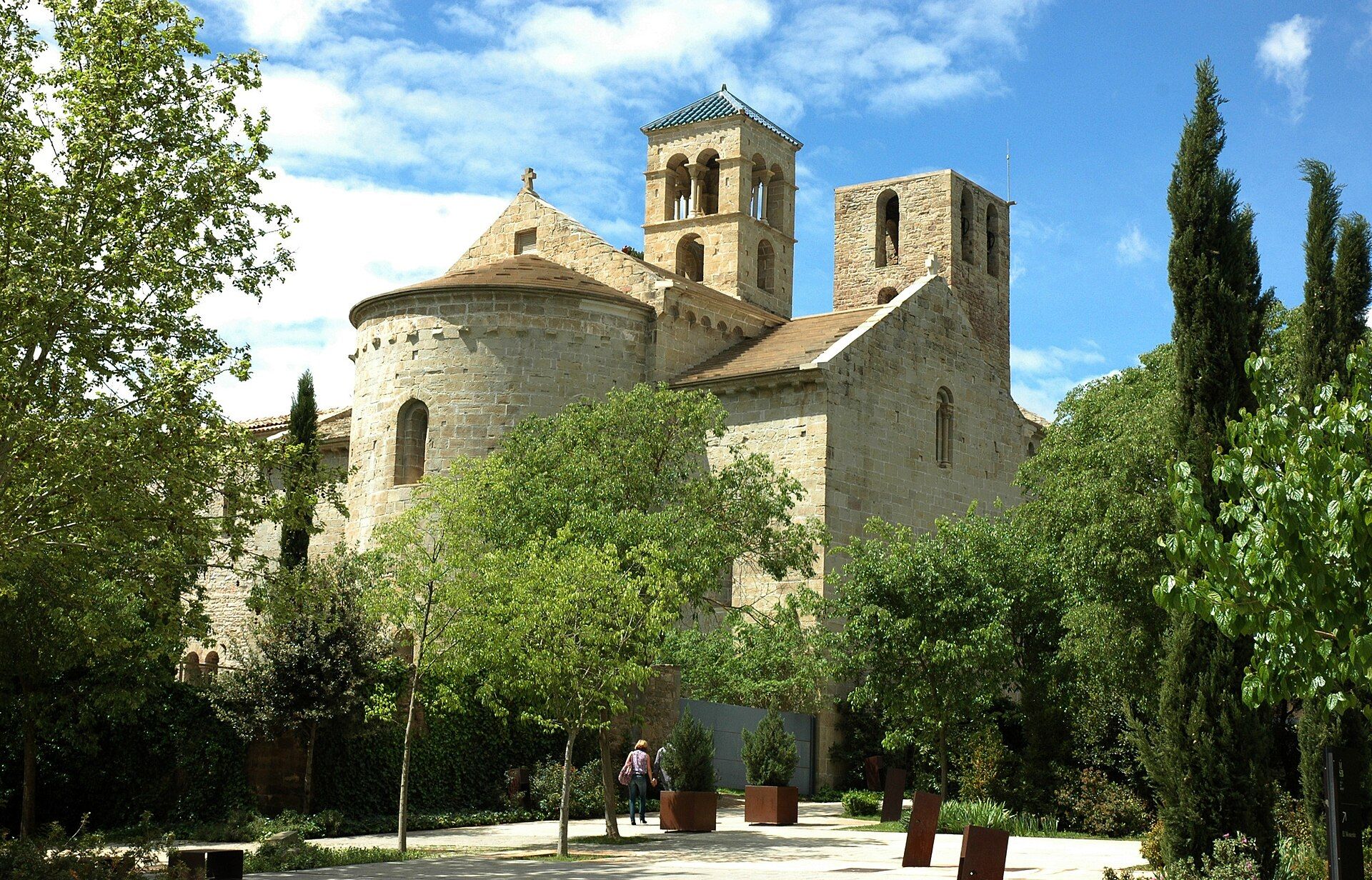 The Romanesque church of Sant Benet de Bages, with its round apse and two square bell towers, seen through trees and cypresses