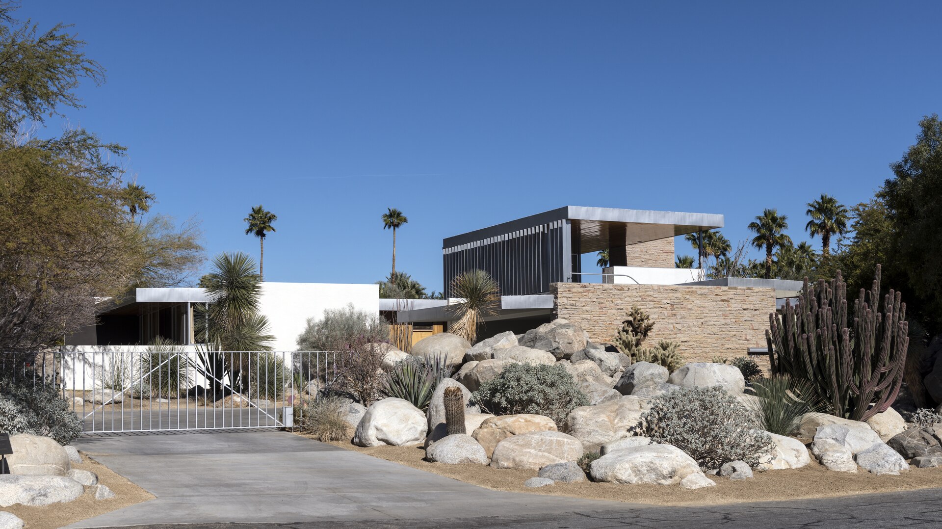 A low flat-roofed desert house behind a gate, with boulders and cactus in the front garden