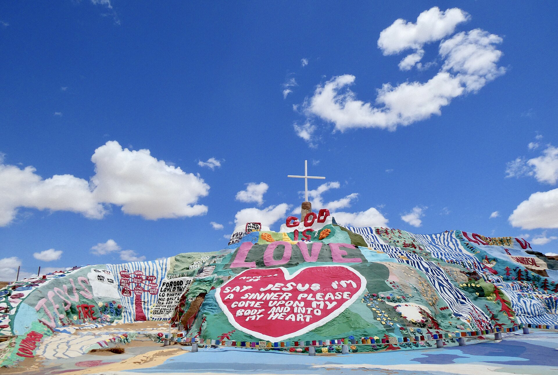A painted adobe hillside covered in bright stripes, flowers and lettering under a blue sky