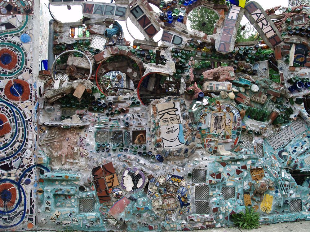 A wall covered in mosaic made from bottles, mirrors, tiles and a bicycle wheel