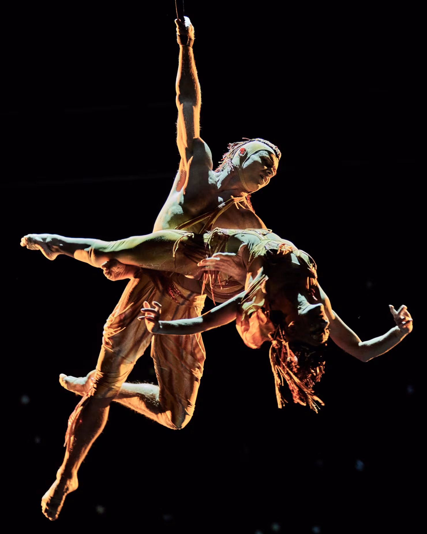 Two aerialists intertwined in mid-air against a black stage in Mystère